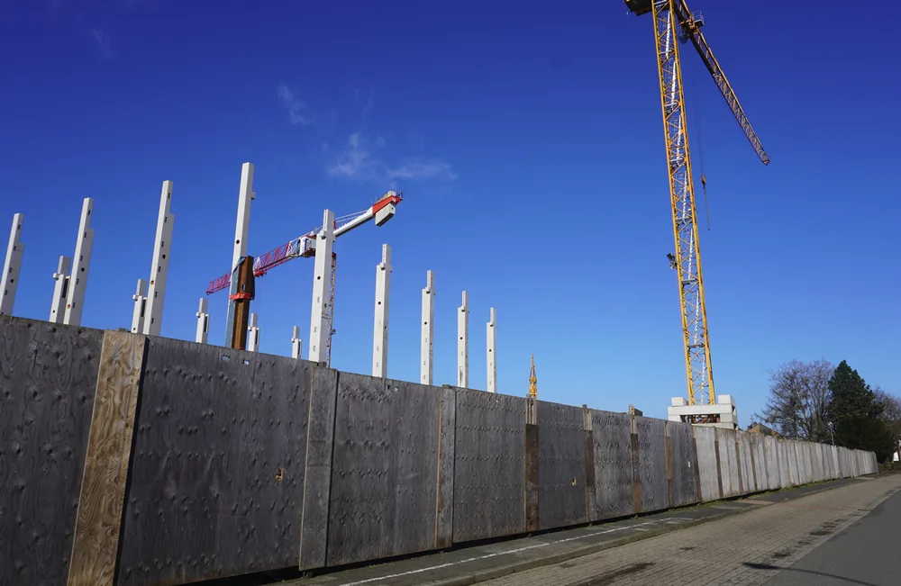 Die hohe Holzwand schützt die Baustelle der neuen Petrus-Canisius-Grundschule in Weeze. Dahinter ragen bereits Kräne und Betonstützen in den Himmel.