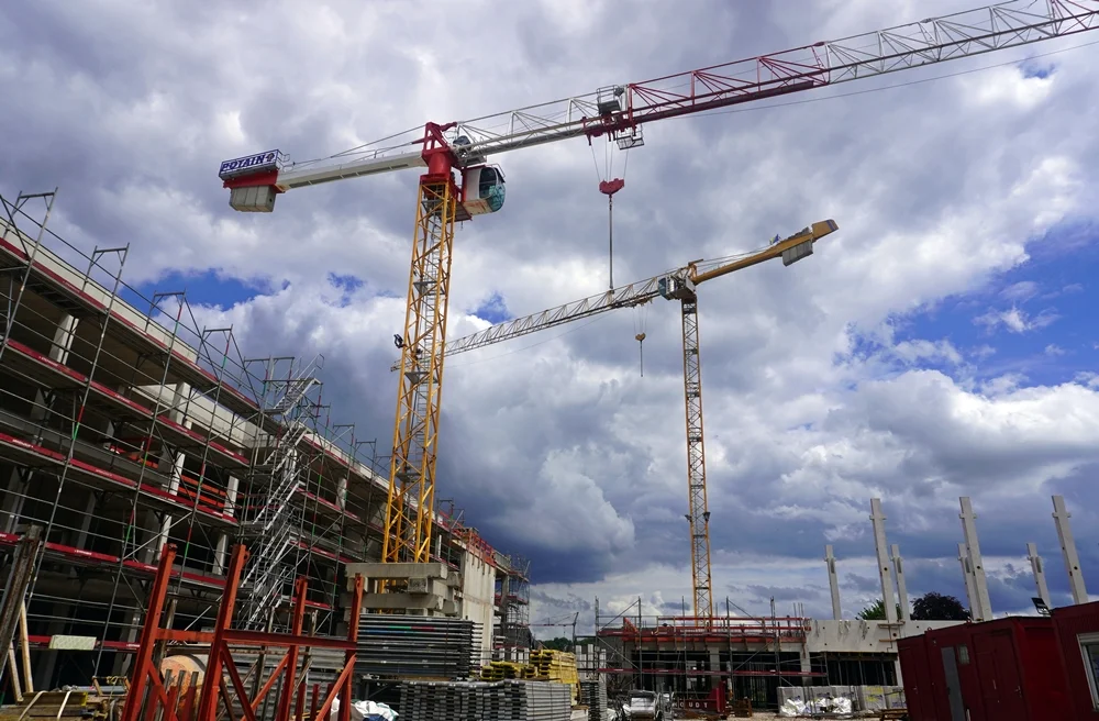 Zwei große Baukräne ragen über die Baustelle der neuen Schule in Weeze. Links ist das eingerüstete Hauptgebäude zu sehen, rechts entstehen weitere Gebäudeteile unter einem dramatischen Wolkenhimmel.