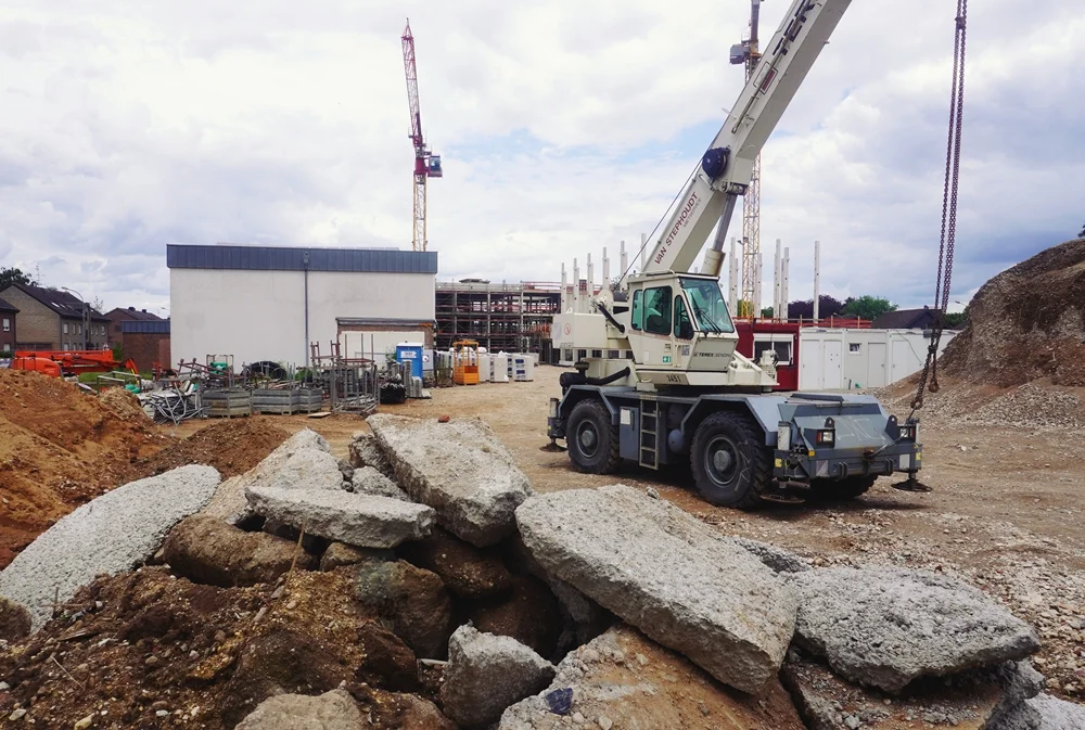 Ein Mobilkran steht auf der Baustelle der neuen Schule in Weeze. Im Vordergrund liegen große Betonreste, während im Hintergrund der Rohbau und mehrere Baukräne zu sehen sind.