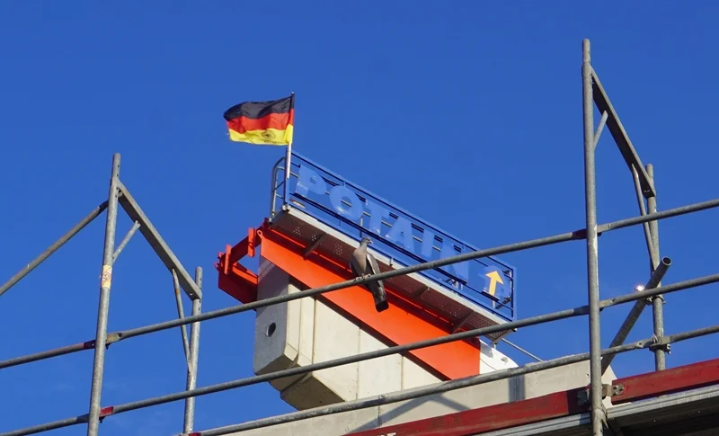 Deutschlandfahne auf einem Baukran während der Fußball-WM-Zeit. Die Fahne weht vor strahlend blauem Himmel über einem Rohbau. Zwischen Gerüststangen ist der Kranaufbau mit Firmenlogo zu sehen. Die Aufnahme vermittelt Baustellenalltag mit einem Hauch von Fußballbegeisterung und nationaler Stimmung.