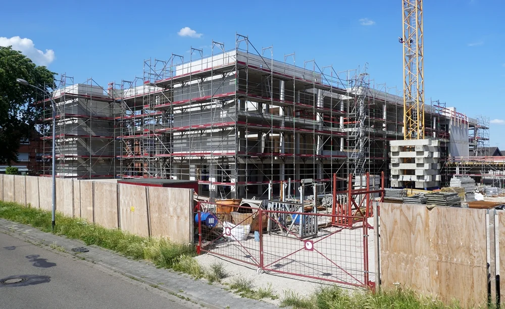 Rohbau der neuen Petrus-Canisius-Grundschule in Weeze Mitte Juni 2026. Das mehrstöckige Schulgebäude ist von Gerüsten umgeben, während auf dem Baustellengelände Baumaterialien und ein Baukran zu sehen sind. Ein Bauzaun trennt die Baustelle von der Straße im Vordergrund. Über dem Neubau spannt sich ein sonniger, wolkenarmer Sommerhimmel.