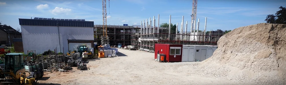Panoramaaufnahme der Baustelle der neuen Petrus-Canisius-Grundschule in Weeze Mitte Juni 2026. Links steht die ehemalige Turnhalle, in der Mitte sind der Rohbau der neuen Mensa mit ihren weißen Betonstützen sowie weitere Schulgebäude im Bau zu sehen. Rechts erhebt sich ein großer Kieshügel, während Baucontainer, Maschinen und Baumaterialien das Baustellengelände prägen.