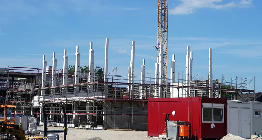 Die im Bau befindliche Mensa der Petrus-Canisius-Grundschule in Weeze. Zahlreiche weiße Betonstützen ragen über das Gerüst hinaus und geben dem Gebäude bereits seine spätere Form. Im Vordergrund stehen rote Baucontainer, während im Hintergrund weitere Teile des Schulneubaus zu erkennen sind. Über der Baustelle erstreckt sich ein heller Sommerhimmel.