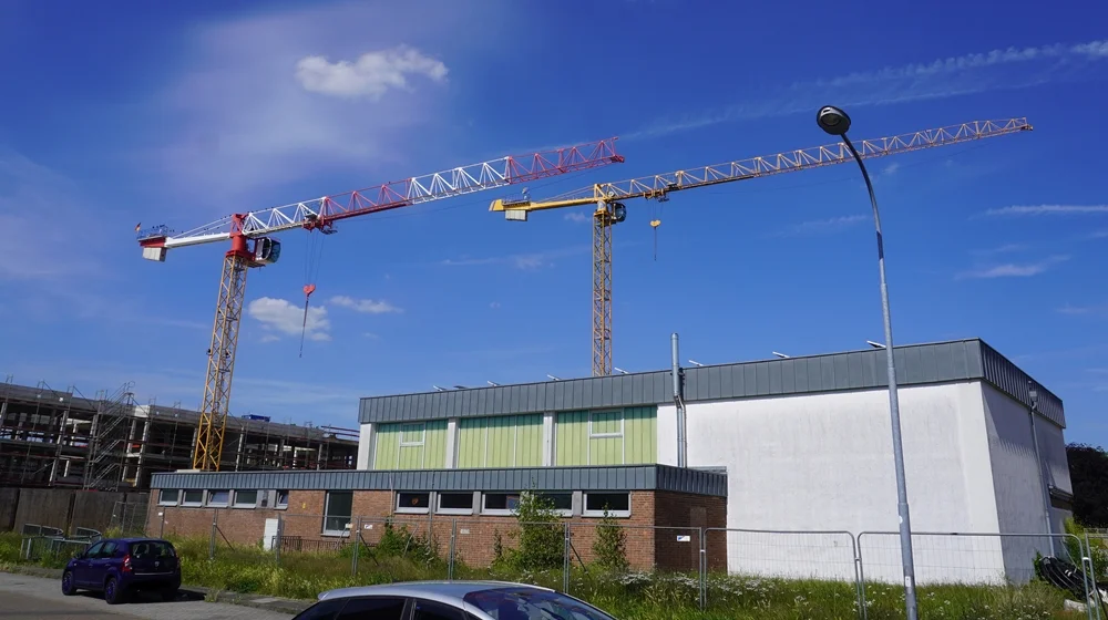 Alte Turnhalle der Johannes-Hauptschule Weeze mit Baukränen und dem Rohbau der neuen Schule im Hintergrund unter blauem Sommerhimmel (2026).