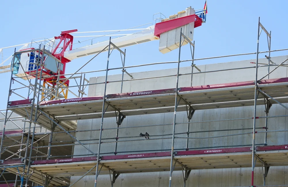 Baugerüst an einem Schulneubau mit Turmdrehkran vor blauem Himmel. Zwei Tauben sitzen im Schatten an der Betonwand.
