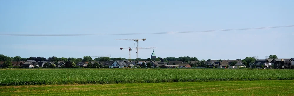 Weitläufige Feldlandschaft vor dem Ortsrand von Weeze. Im Hintergrund sind Häuser, ein Kirchturm und die Kräne der Schulbaustelle zu sehen.