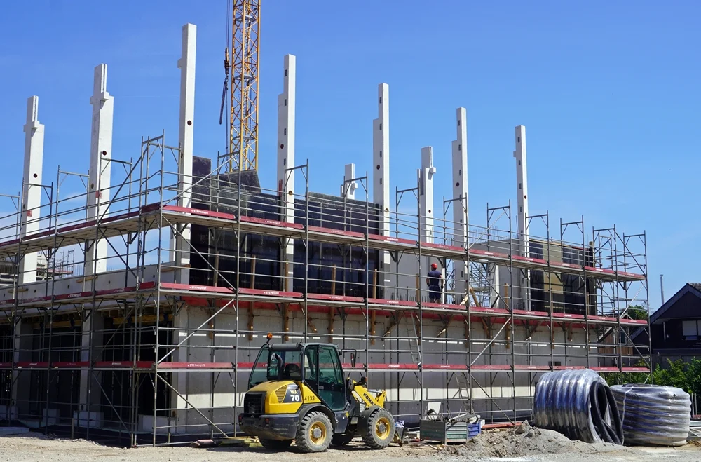 Neubau der Petrus-Canisius-Grundschule in Weeze: Gerüste umgeben die zweite Etage über der Mensa, während Betonstützen und Wandschalungen für die spätere Sporthalle montiert werden. Im Vordergrund steht ein Radlader auf der Baustelle.