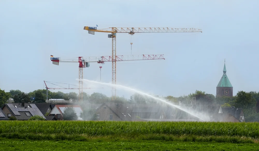 Beregnungsanlage bewässert ein Feld vor der Baustelle der neuen Petrus-Canisius-Grundschule in Weeze. Im Hintergrund sind Baukräne und der Kirchturm der St.-Cyriakus-Kirche zu sehen.