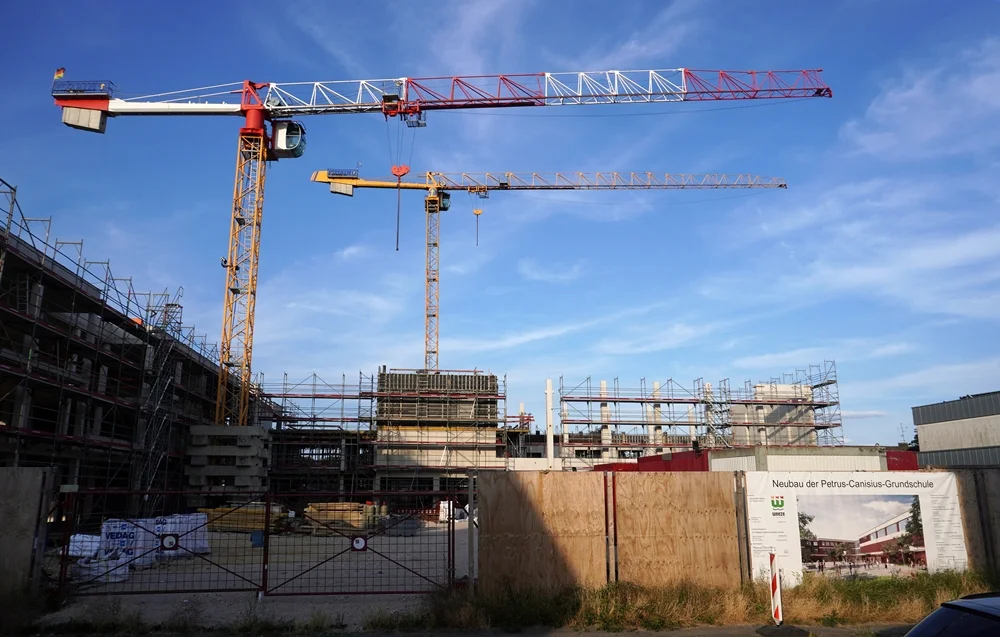 Übersichtsaufnahme der Baustelle der neuen Petrus-Canisius-Grundschule in Weeze mit zwei Baukränen, dem Rohbau und der Bautafel am Bauzaun.